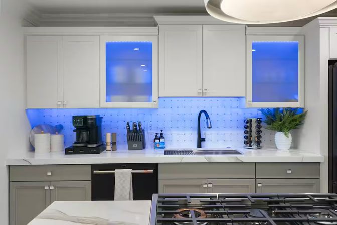 Sleek modern white kitchen with blue LED under-cabinet lighting, marble countertops, black pull-down faucet, coffee maker, knife block, potted fern, and gas cooktop in foreground.