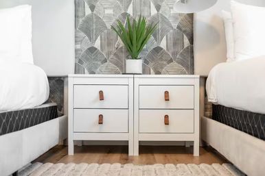 Modern bedroom interior with twin beds flanking matching white nightstands with leather strap pulls, geometric gray wallpaper, and a small green potted plant on a light wood floor.