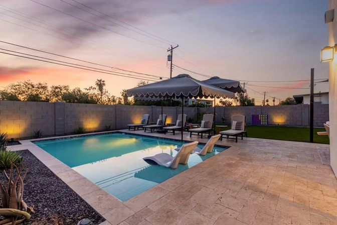 Sunset-lit suburban backyard pool with tiled patio, two in-pool loungers, chaise lounges under scalloped umbrellas and soft wall lighting.
