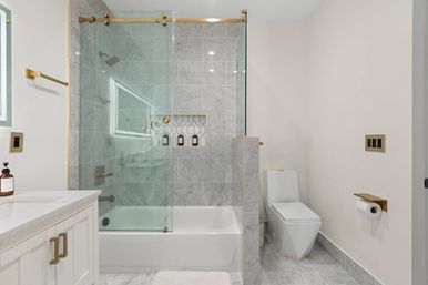 Spa-like modern marble bathroom with tub-shower combo, frameless glass sliding door and brass hardware, tiled shower niche with bottles, white vanity with quartz countertop, and sleek one-piece toilet with gold toilet-paper holder.
