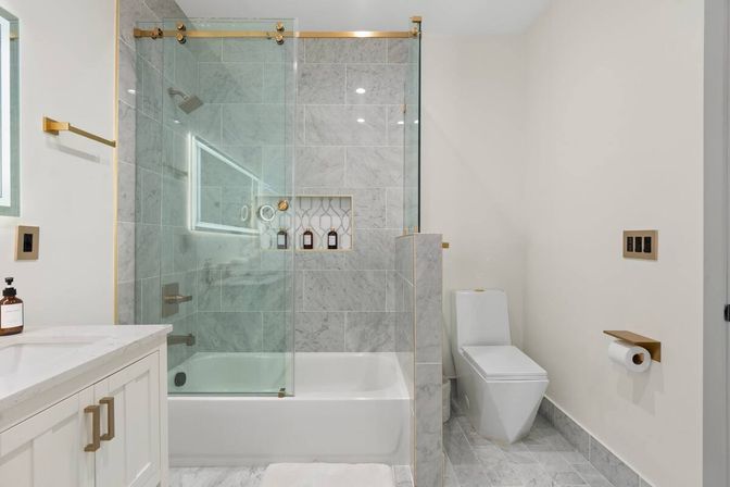 Spa-like modern marble bathroom with tub-shower combo, frameless glass sliding door and brass hardware, tiled shower niche with bottles, white vanity with quartz countertop, and sleek one-piece toilet with gold toilet-paper holder.