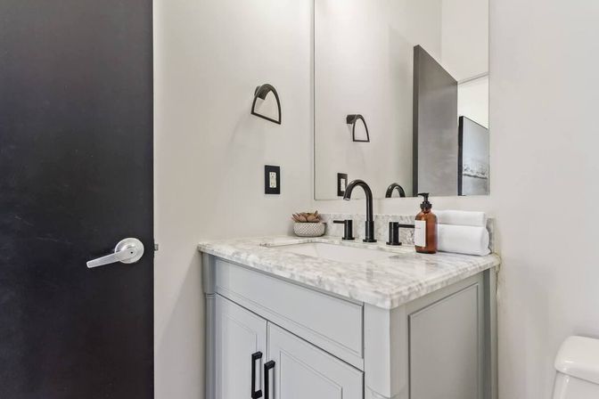 Contemporary powder room with light-gray vanity and marble countertop, matte-black faucet and hardware, rectangular mirror, amber soap dispenser, rolled white towel and small succulent on the counter.