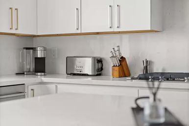 Bright modern white kitchen countertop with stainless steel toaster, single-serve coffee maker, wooden knife block and gas cooktop beneath white cabinets.