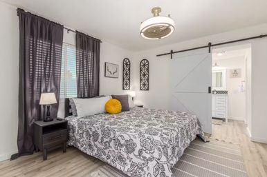 Contemporary master bedroom with gray floral duvet, bright yellow round accent pillow, dark curtains, nightstand lamp, sliding white barn door to en-suite bathroom and light wood floors.