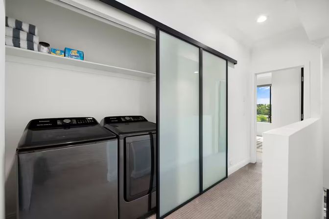 Modern hallway laundry closet with black washer and dryer behind frosted sliding glass doors, open shelf with towels and detergent, bright white contemporary home interior with windowed room beyond