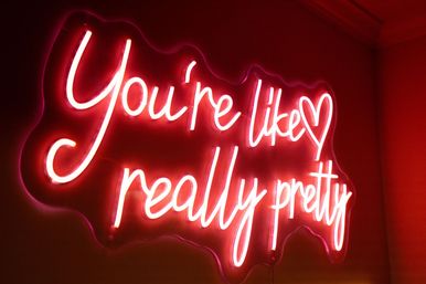 Pink neon cursive wall sign reading 'You're like really pretty' with heart accent, glowing red-pink ambient light and modern interior decor