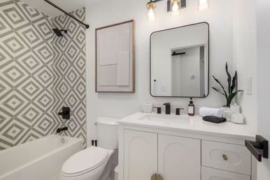 Bright modern white bathroom with bathtub and gray geometric tile shower, white vanity with sleek black faucets and rounded-square mirror, toilet and small potted plant.