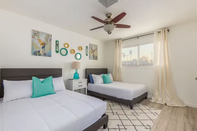 Sunlit modern guest bedroom with two gray-framed beds in white linens and teal pillows, white nightstand with turquoise lamp, ceiling fan, geometric rug, wall art, and palm trees visible through the window.
