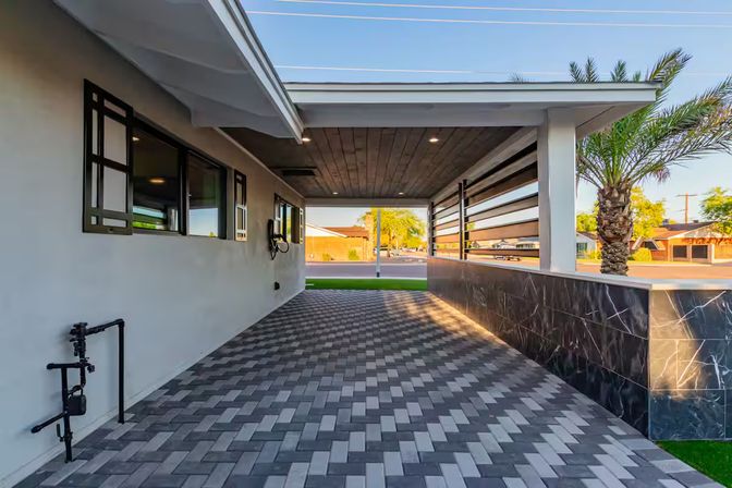 Contemporary covered carport with herringbone gray paver driveway, wood-paneled ceiling with recessed lights, marble-look half wall and palm tree opening to a sunny suburban street.