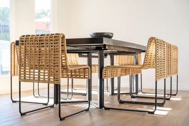 Sunlit modern dining room interior with a black metal table and six woven rattan chairs on light wood flooring near tall windows