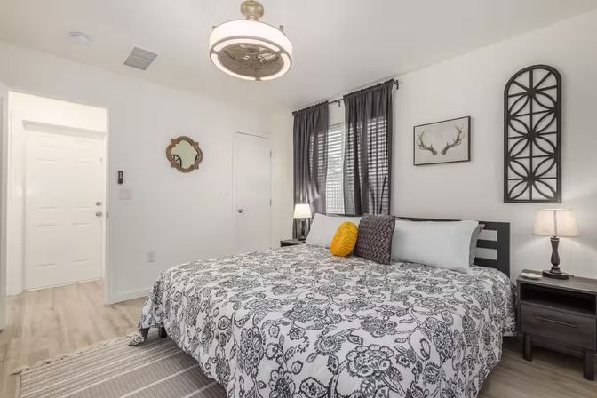 Cozy modern master bedroom with king bed, black-and-white paisley duvet, yellow accent pillow, gray curtains, bedside lamps and decorative wall art.
