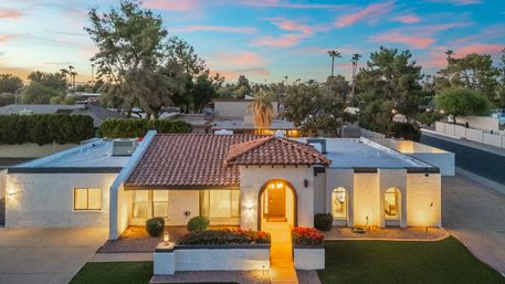 Single-story Southwestern stucco home with red tile roof and arched entry, warm exterior lighting, manicured front garden and palm trees under a colorful sunset sky in a suburban neighborhood.