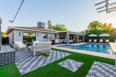 Modern backyard oasis with a rectangular swimming pool, paved patio and walkways, wicker lounge seating and hanging egg chairs, poolside loungers with umbrellas, and a covered patio attached to a single-story home.