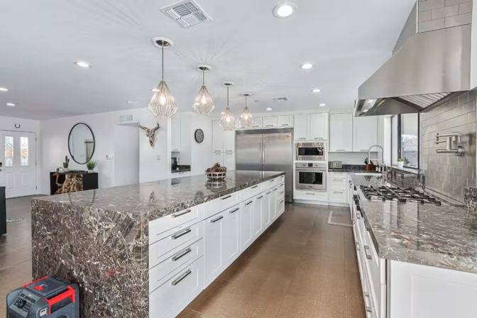 Bright open-concept modern kitchen with a long dark marble island, white cabinetry, stainless steel appliances, pendant lights, and wood-look floors.