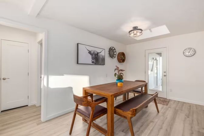 Sunlit modern dining room with mid-century wooden table, matching bench and chairs, blue-and-yellow potted plant centerpiece, black-and-white longhorn wall art, round shelves, skylight and glass door on light wood floors.