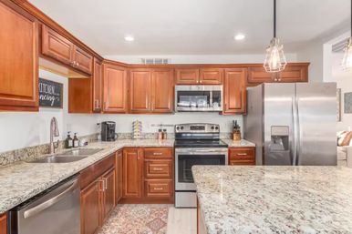 Bright modern kitchen interior with cherry wood cabinets, speckled granite countertops and island, stainless steel refrigerator, oven and dishwasher, pendant lights and undermount sink.