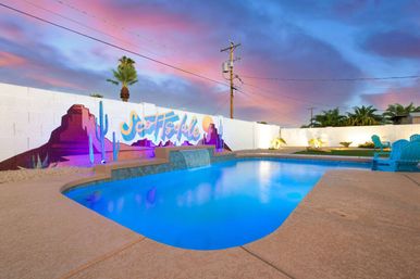 Backyard swimming pool at dusk with a colorful Scottsdale desert mural on a white wall, illuminated spillover waterfall, palm trees and turquoise Adirondack chairs