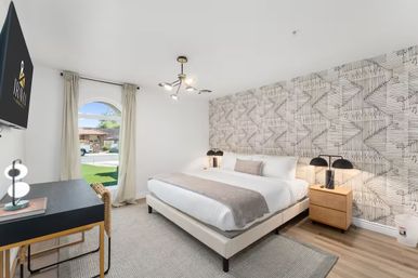 Bright modern bedroom with king bed, geometric patterned accent wall, wood nightstands with black lamps, a small desk and chair, arched window showing a suburban street view, and hardwood floors.