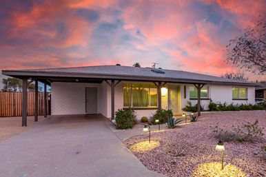 Twilight single-story ranch home with covered carport, xeriscape rock front yard and glowing pathway lights under a pink sunset sky.