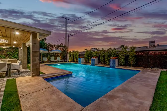 Residential backyard pool at sunset with illuminated blue water, modern covered patio dining area, lounge chairs with umbrellas, and landscaped privacy hedges