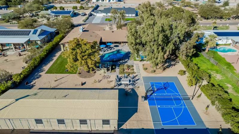 Aerial drone view of a sunny suburban property with a kidney-shaped swimming pool, patio umbrellas and seating, mature trees, a vivid blue outdoor basketball court, a large metal-roofed outbuilding, and neighboring homes with solar panels and desert-style landscaping.