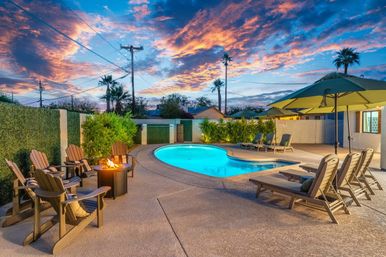 Twilight resort-style backyard with glowing blue kidney-shaped pool, lounge chairs and umbrellas, Adirondack chairs around a fire pit, palm trees and dramatic pink-orange sunset sky.