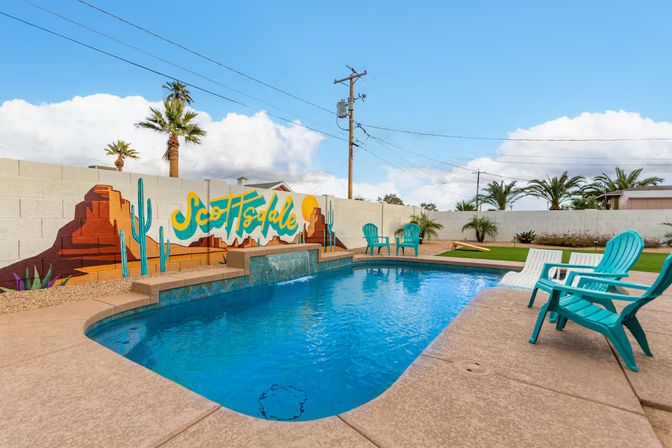 Sunny Scottsdale backyard with a blue swimming pool, colorful desert mural on a block wall reading "Scottsdale", turquoise Adirondack chairs, palm trees and clear sky.