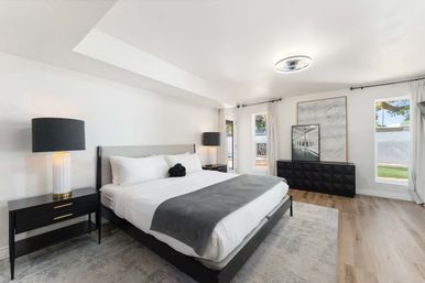 Sunlit contemporary master bedroom with king-size bed, white linens and gray throw, black nightstands and lamps, geometric black dresser, large windows with curtains, hardwood floors and neutral minimalist decor.