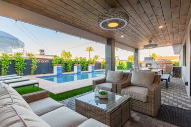 Sunny backyard oasis with a covered wooden-ceiling patio featuring wicker sofa and armchairs, glass-top coffee table, dining area and string lights overlooking a rectangular pool with tiled water features, lounge chairs and palm trees.