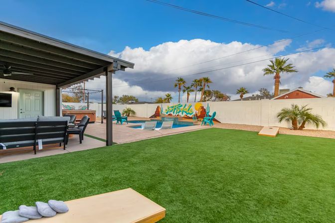 Sunny backyard with small pool and in-pool loungers, covered patio seating, artificial turf lawn, cornhole boards, palm trees and a colorful desert mural