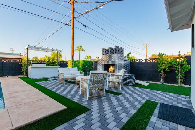 Modern sunny backyard patio with gray-brick outdoor fireplace, wicker lounge seating on patterned pavers, pergola bar, pool edge and palm trees.