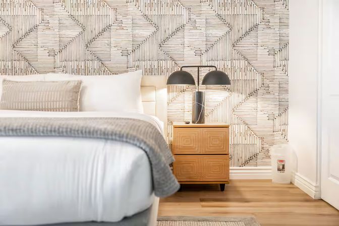 Contemporary bedroom interior with white bedding, gray throw blanket, geometric textured wallpaper, wooden nightstand and double‑dome lamp on hardwood floor.