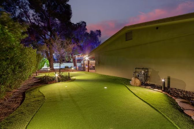 Twilight suburban backyard putting green with smooth artificial turf, low landscape lighting, stone edging, large trees and a lit patio pergola for evening practice