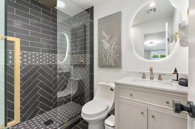 Sleek modern bathroom with glass-enclosed shower showcasing black herringbone and geometric tiles, white toilet and vanity with gold faucets, and a round backlit mirror.