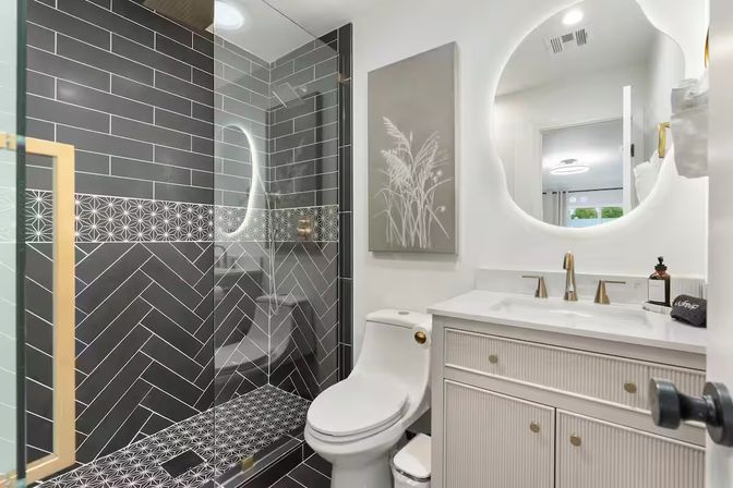 Sleek modern bathroom with glass-enclosed shower showcasing black herringbone and geometric tiles, white toilet and vanity with gold faucets, and a round backlit mirror.