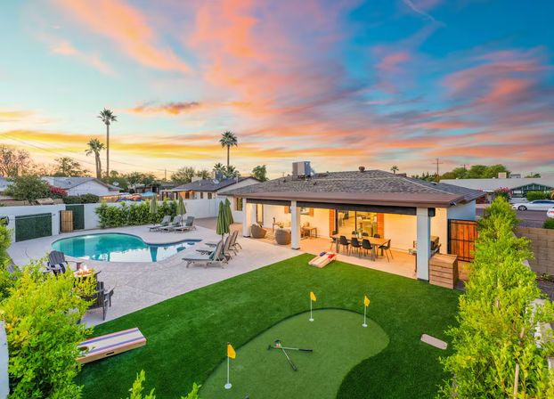 Suburban backyard at sunset with a kidney-shaped pool, artificial putting green, covered patio dining area, lounge chairs and palm trees under a colorful pink-orange sky.