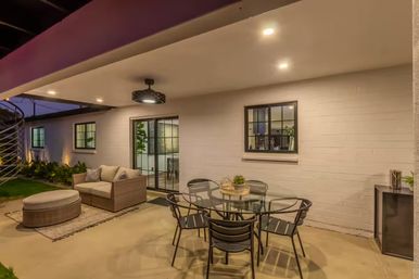 Cozy modern covered backyard patio at dusk with round glass dining table and six metal chairs, wicker loveseat and ottoman on a rug, recessed lights and black pendant by a sliding glass door and white brick wall.