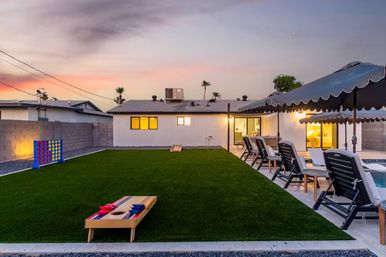 Sunset backyard with green artificial lawn, cornhole boards and oversized Connect Four, row of lounge chairs under scalloped canopies beside a pool and a warmly lit single‑story home with palm trees