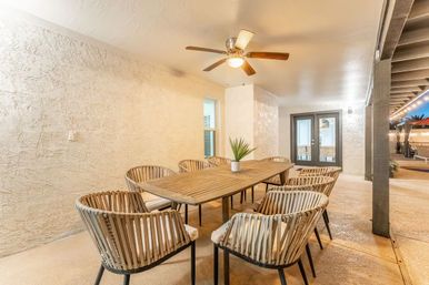 Covered backyard patio dining area with a long wooden table, eight woven rattan chairs, ceiling fan overhead and a small potted plant centerpiece, cozy evening-ready outdoor entertaining space with glass doors leading inside.