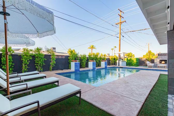 Sunlit suburban backyard with rectangular swimming pool, blue-tiled water features, concrete deck, row of white chaise lounges under fringed umbrellas, manicured hedges, palm trees and an outdoor fireplace.