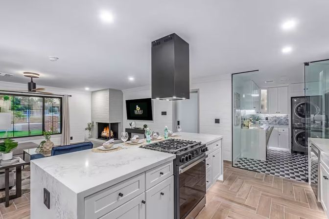 Bright open-plan modern kitchen and living area with white marble island, gas range and black vent hood, cozy fireplace, large picture window, glass-enclosed laundry with stacked washer-dryer, and wood-look tile flooring.