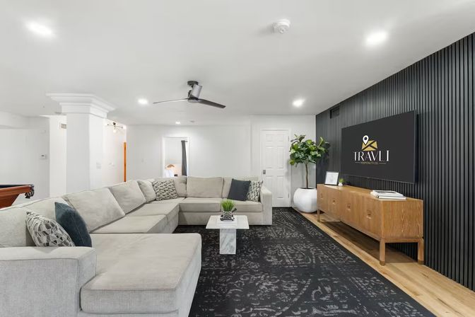 Cozy chic modern basement living room featuring a large light-gray sectional, marble coffee table, black patterned rug, wood media console and wall-mounted TV on a black slatted accent wall, ceiling fan and potted plant.