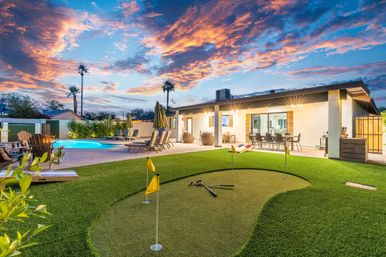 Modern single-story backyard at sunset with illuminated pool, putting green with flags, outdoor dining patio and palm trees