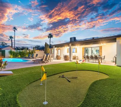 Modern single-story backyard at sunset with illuminated pool, putting green with flags, outdoor dining patio and palm trees