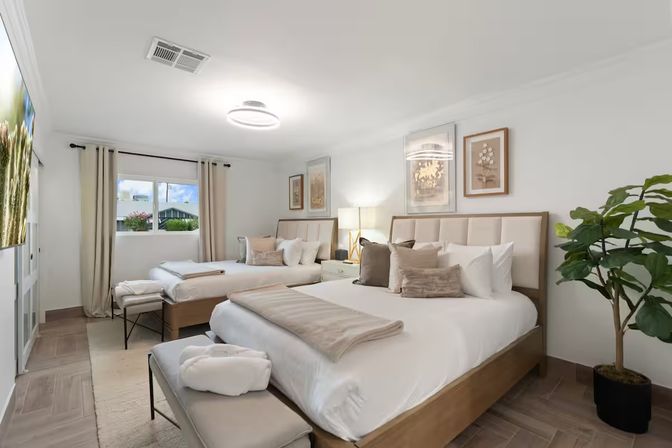 Bright modern guest bedroom showcasing two comfy queen beds with neutral linens and throw pillows, light wood bed frames and bench, beige curtains over a large window, framed wall art, potted plant, and wood-look tile flooring.