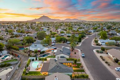Aerial view of a suburban neighborhood at sunset — single-story homes with backyard pools, palm trees and desert landscaping, wide streets leading toward distant silhouetted mountains under a colorful sky.