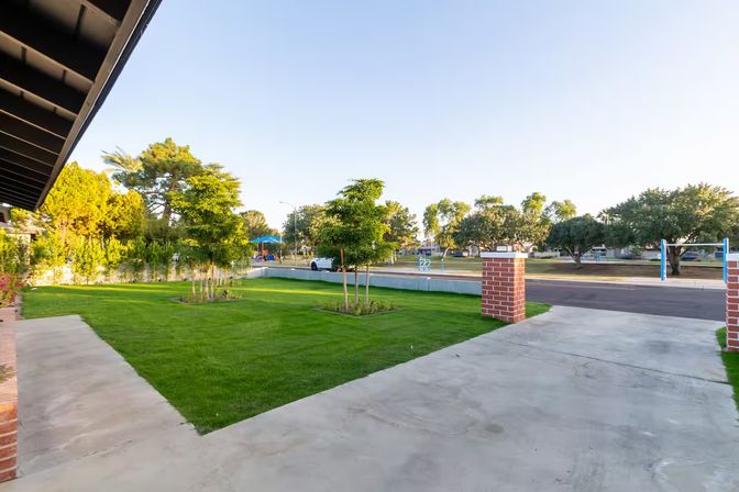 Sunlit suburban front yard with neatly mowed green lawn, young trees, concrete driveway framed by brick pillars, sidewalk and a neighborhood park with playground and mature trees under a clear blue sky