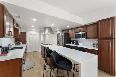 Sleek modern kitchen with a long white quartz island and black bar stools, dark wood cabinets, stainless steel refrigerator, microwave and cooktop with a stainless hood, light hardwood floors and recessed lighting.