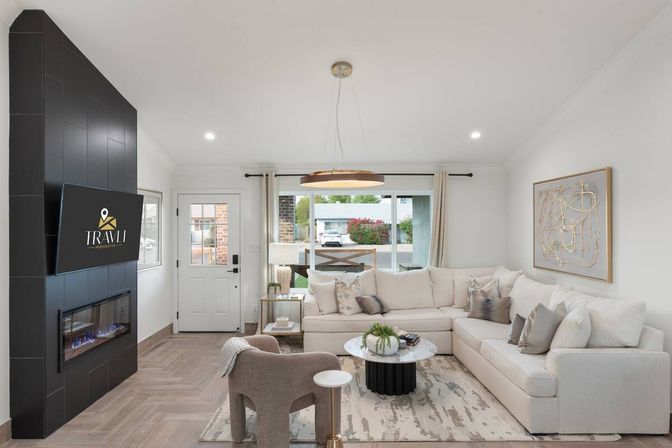 Cozy, bright suburban living room with a neutral palette — beige L-shaped sectional, toss pillows, round marble coffee table, patterned rug, gray accent chair, pendant light, sleek black-tiled wall with electric fireplace and wall-mounted TV, and a front door and large window letting in natural light and a street view.