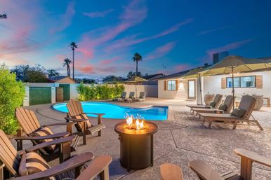 Sunset backyard with glowing kidney-shaped pool, round fire pit surrounded by Adirondack chairs, cushioned loungers under umbrellas, palm trees and single-story patio home